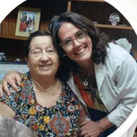 Una profesional de Ámbar, de guardapolvo, abraza sonriendo a una mujer mayor en el living de su casa.