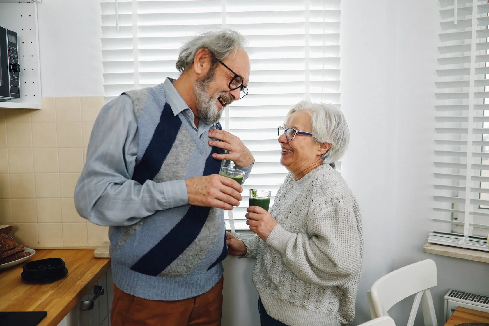 Una pareja mayor se ríe en la cocina de su casa mientras comparte un vaso de jugo verde.